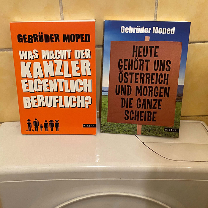 Zwei Bücher von Gebrüder Moped auf einer WC-Spülung: links das orangefarbene Cover „Was macht der Kanzler eigentlich beruflich?“, rechts das Buch „Heute gehört uns Österreich und morgen die ganze Scheibe“
