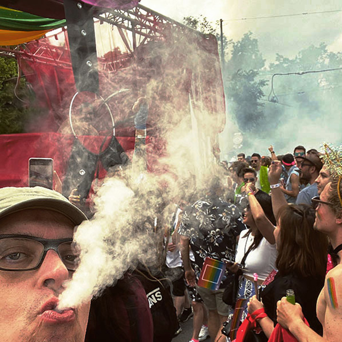 Franz Moped auf der Vienna Pride vor einem Wagen, der als Effekt Dampf ablässt. Auch aus dem Mund von Franz Moped kommt Dampf einer E-Zigarette.