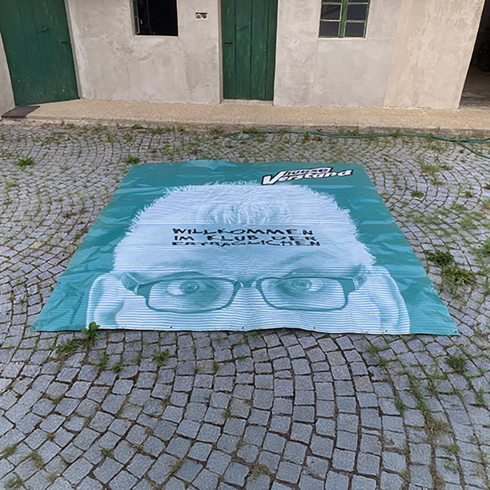 Bühnenbanner der früheren Band „Houseverstand“ mit dem Gesicht von Franz Moped und dem Schriftzug „Willkommen im Klub der Erträglichen“, ausgebreitet im gepflasterten Innenhof eines alten Bauernhauses