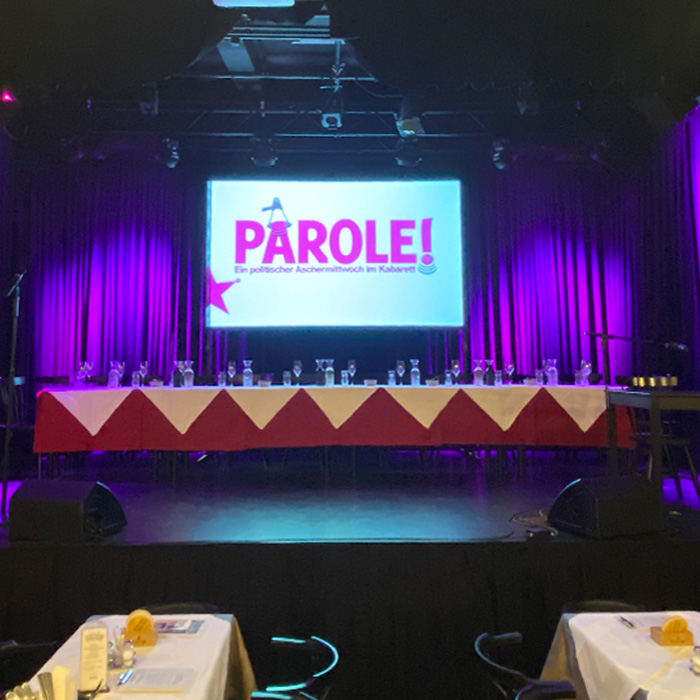 Der für 12 Personen gedeckte, lange Tisch auf der Bühne der Kulisse Wien vor der Veranstaltung der Gebrüder Moped zum politischen Aschermittwoch. Hinten auf die Bühne projiziert: "Parole!"