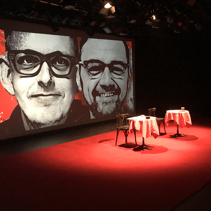 Leere Bühne der Produktion "Heute gehört uns Österreich und morgen die ganze Scheibe" der Gebrüder Moped 2018 im Rabenhof Theater Wien. Die Bühne in Rot. Zwei Tische, zwei Sesseln, dahinter projiziert über die ganze Rückenwand der Bühne: die Gesichter von Franz und Martin Moped